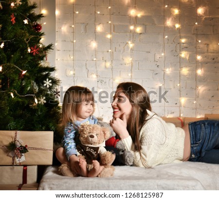 young mother and a little girl daughter are sitting at home on the sofa near the New Year tree. lights of garlands. comfort and warmth, homely atmosphere, preparing for the new year
