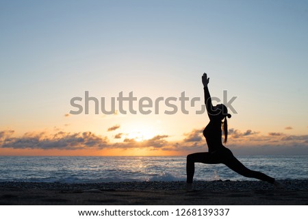 Human silhouette doing yoga on the beach in front of rising sun, woman stands in warrior pose, hands up. Wellness and activity concept