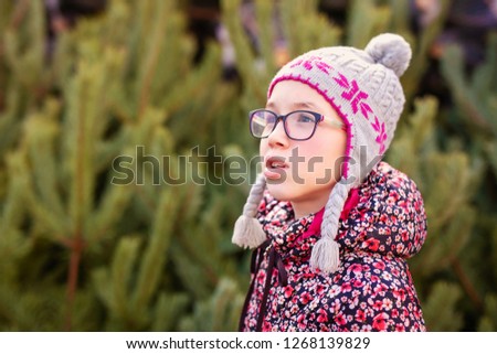Girl with big glasses and gray and purple hat looking at green christmas tree.