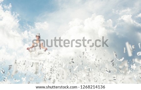 Woman in white clothing keeping eyes closed and looking concentrated while meditating among flying letters in the air with cloudy skyscape on background.