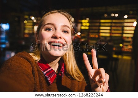 Hipster female blogger making photo on front camera at her smartphone device sitting in loft cafe or pub interior in evening.Excited blond woman taking selfie on mobile phone for social networks.