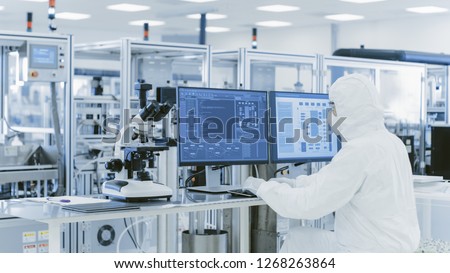 In Laboratory Over the Shoulder View of Scientist in Protective Clothes Doing Research on a Personal Computer. Modern Manufactory Producing Semiconductors and Pharmaceutical Items.
