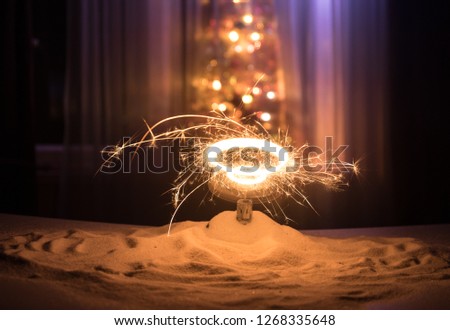 Glittering burning sparkler on snow with blurred Christmas tree on dark background. New Year Holiday concept with empty space for your text. Selective focus