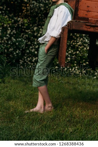a boy in a white shirt and green pants, a green sweater around his neck, leaning his elbows on the old truck cousin, barefoot on the grass in the summer. village, farmer,on farm. hands in pockets