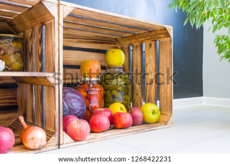 Apples, cabbage and pickled cucumbers and tomatoes in jars are in a wooden box on background of black wall in kitchen. Aesthetic open storage of products or stocks of food in context of modern kitchen