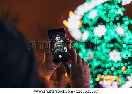Close-up hands of young woman in warm gloves holding mobile phone photographing glowing green lights of Christmas tree in city at night.