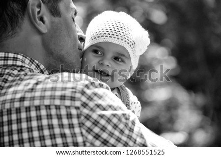 Father kissing and holding small daughter baby girl cute hazel-eyed kid tiny little child wearing white flower beanie hat outdoor on sunny summer day on blurred green background, horizontal picture 