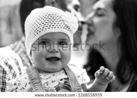 Girl cute smiling baby hazel-eyed kid tiny little child wearing white flower beanie hat in arms of kissing parents mother and father family summer day on blurred green background, horizontal picture 