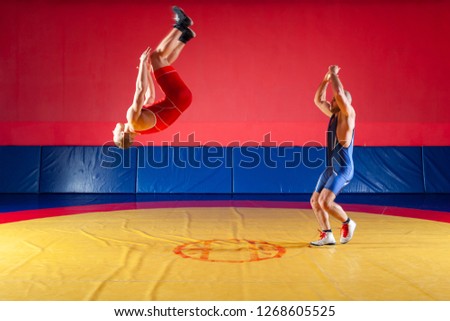 Two strong wrestlers in blue and red tights make flips and jumps on the yellow wrestling track in the gym. A young man makes a grab.