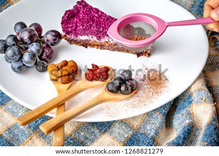 Raw cake on plate with seeds and fruit ingredients on wooden spoon. Vegan cake with grapes and berries. Healthy cake with goji seeds and nuts.