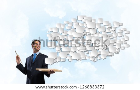 Happy and young businessman in black suit holding paintbrush in his hand and smiling while standing with messages in form of cloud against blue cloudy skyscape view on background.