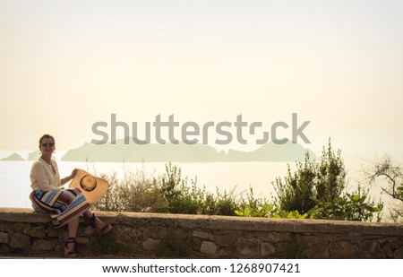Young woman sitting on a wall at sunset in front of the sea on Ponza island coast. Fashion white shirt, colorful skirt, large hat, flowers bouquet.