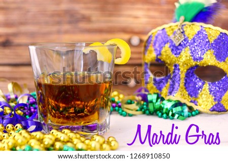 A sazerac cocktail with a lemon twist in a rocks glass on a wooden table. Mardi gras decorations around. Wooden background. Text added.
