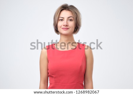 Charismatic attractive caucasian woman in red dress, smiling cheerfully while standing against gray background, posing for id photo
