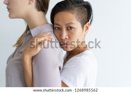 Asian woman embracing girlfriend from behind and looking at camera. Multiethnic homosexual couple. Lesbian couple concept. Isolated cropped side view on white background.