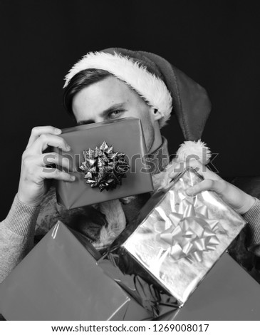 Man holds christmas gifts. Santa hiding behind presents on black background. New Year and Christmas concept. Guy with smiling face in red hat with red and silver gift boxes.