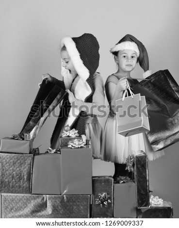 Children with surprised faces stand on blue background. Childhood and sale concept. Sisters in Christmas hats with gift boxes open presents and shopping bags. Girls celebrate Christmas