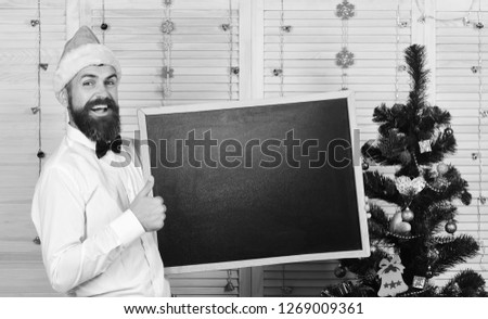 Guy near Christmas tree on wooden wall background. Man with beard holds blank blackboard, copy space. Celebration and New Year mood concept. Santa Claus in red hat with happy face shows thumbs up
