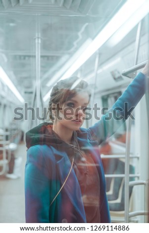 Young beautiful girl with brown hairs and bright make up, dressed in blue coat, black hoodie, sneakers and checkered trousers posing in metro carriage. Posing behind the glass
