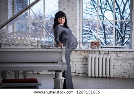 Pregnant woman in the gray dress stands near white piano and the window, winter landscape outside