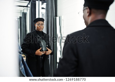 Stylish casual african american man at black beret and overcoat with waist bag at fitting room clothes store, looking on mirror.