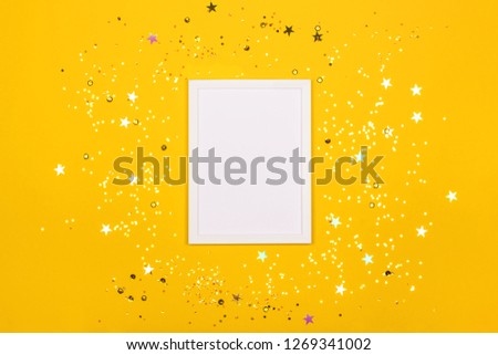 Festive background with blank white photo frame on yellow with scattered confetti. Mock up for photo or text. Top view. Flat lay style.