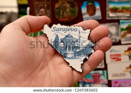 A hand holding a magnet shaped in a form of Bosnia and Herzegovina with pictured famous fountain Sebilj Brunnen in Sarajevo, the capital of Bosnia-Herzegovina