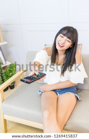 An Asian woman sits on the couch watching TV. Her hand is holding a TV remote control. The TV program is wonderful.
