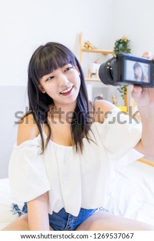 An Asian woman sits on the bed and takes a selfie by camera. This is lifestyle.
