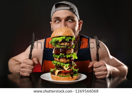 Young hungry man with cutlery eating huge burger on black background