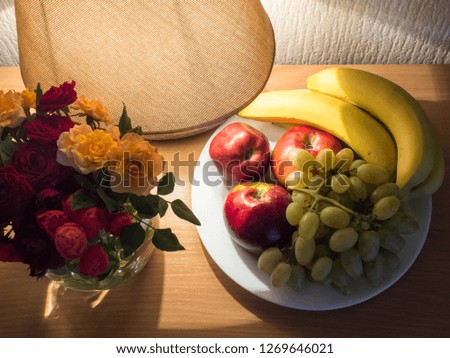 A plate of apples, bananas and grapes next to a table lamp and a vase of roses. Photo in warm colors.