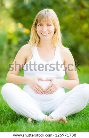 Happy pregnant woman sitting on green grass and holding hands on belly in shape of heart