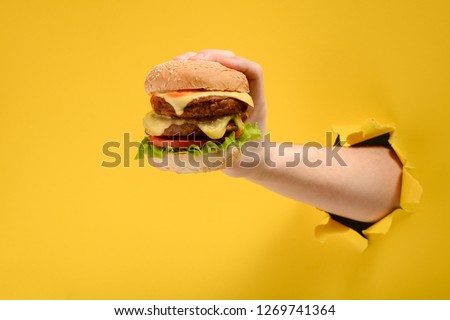 Hand holding a big burger through a hole in torn yellow paper wall. Special offer and cheap price on fast food.