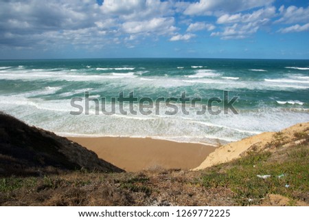 View of the stormy sea from the top of the cliff. The cliff is visible in the picture. Bright blue, slightly overcast sky, beautiful foamy waves.