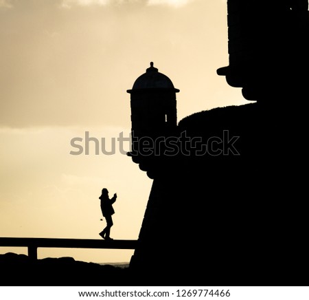 Silhouette of a senior man approaching a lighthouse and taking a shot with his camera. Vertical copy space. Sunset, cloudy sky.