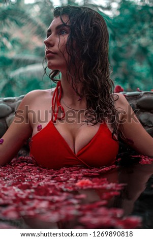 spa tab for relaxion. Aromatherapy treatment. Red flowers floating in the pool. A girl sits in a flower pool in the jungle among the leaves. Brunette with nature big breasts in red swimsuit