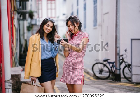 2 young women friends have a video call with friends on a smartphone as they stand next to one another on a street during the day. They are both Asian but diverse, one is Chinese the other Indian.