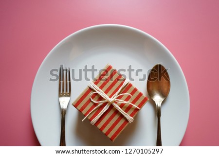 New Year celebrations or Nutritious foods for good health theme red and brown striped gift box with white tied rope in a bow on white plate with fork and spoon over pink background. (space for text)