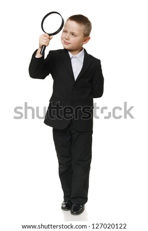 Full length of a schoolboy in black suit looking through the magnifying glass, over white background