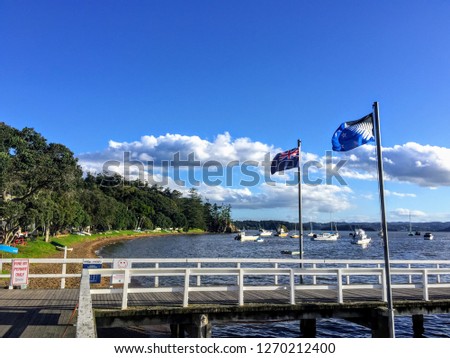 The view of the pier and Kororareka Bay in Russell, New Zealand.  Many boats are anchored in the bay.  The new and old New Zealand flags flap in the wind.  It is a beautiful summer day with blue sky.
