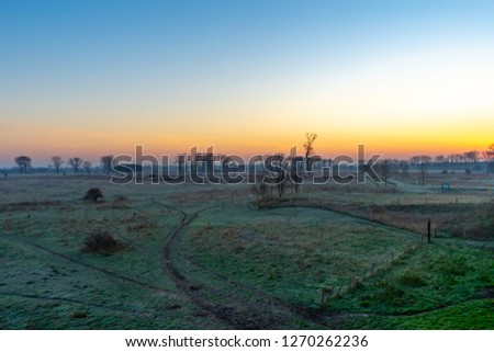 Several paths run through a Dutch river delta landscape on the Belgian border. The rising sun colors the sky orange and blue.