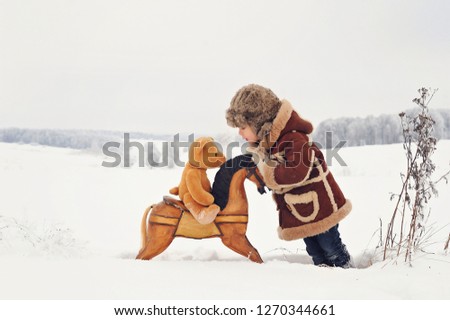 Child in winter. A child playing outside in the snow. The boy in a brown sheepskin coat, with a wooden rocking horse, stands in the snow, in the background a beautiful snowy winter.