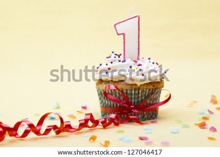 Close-up shot of a cupcake with number 1 candle tied with red streamer over plain yellow background.