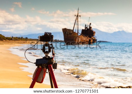 Professional camera on tripod taking picture film video from coastline with rusty shipwreck