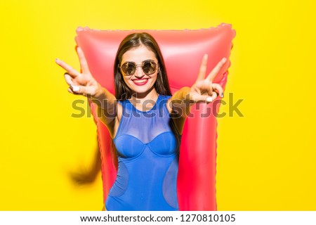 Young attractive woman with peace gesture in blue bikini and sunglasses posing with pink inflatable mattress on yellow background