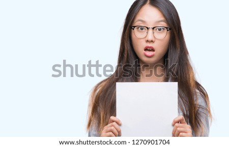 Young asian woman holding blank paper over isolated background scared in shock with a surprise face, afraid and excited with fear expression