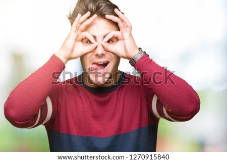 Young handsome man over isolated background doing ok gesture like binoculars sticking tongue out, eyes looking through fingers. Crazy expression.