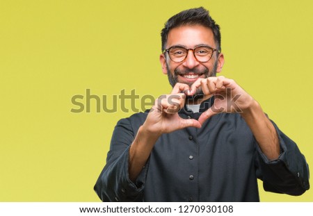 Adult hispanic catholic priest man over isolated background smiling in love showing heart symbol and shape with hands. Romantic concept.