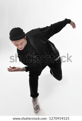 Young freestyle dancer dressed in black jeans, sweatshirt, hat and gray sneakers is dancing in the studio on the white background