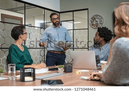 Confident mature businessman giving a presentation to his team in office. Business brief with annual goals with employees and mature boss in meeting room. Multiethnic leader training business people. Royalty-Free Stock Photo #1271048461